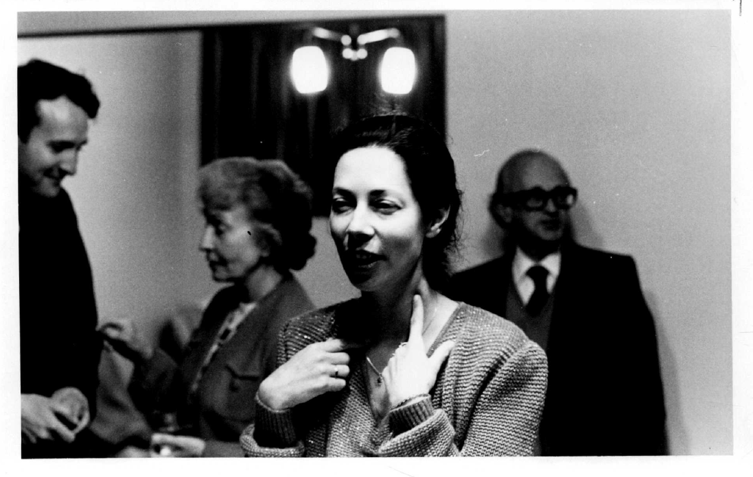 A black and white image of a woman speaking to someone out of frame with people talking in the background
